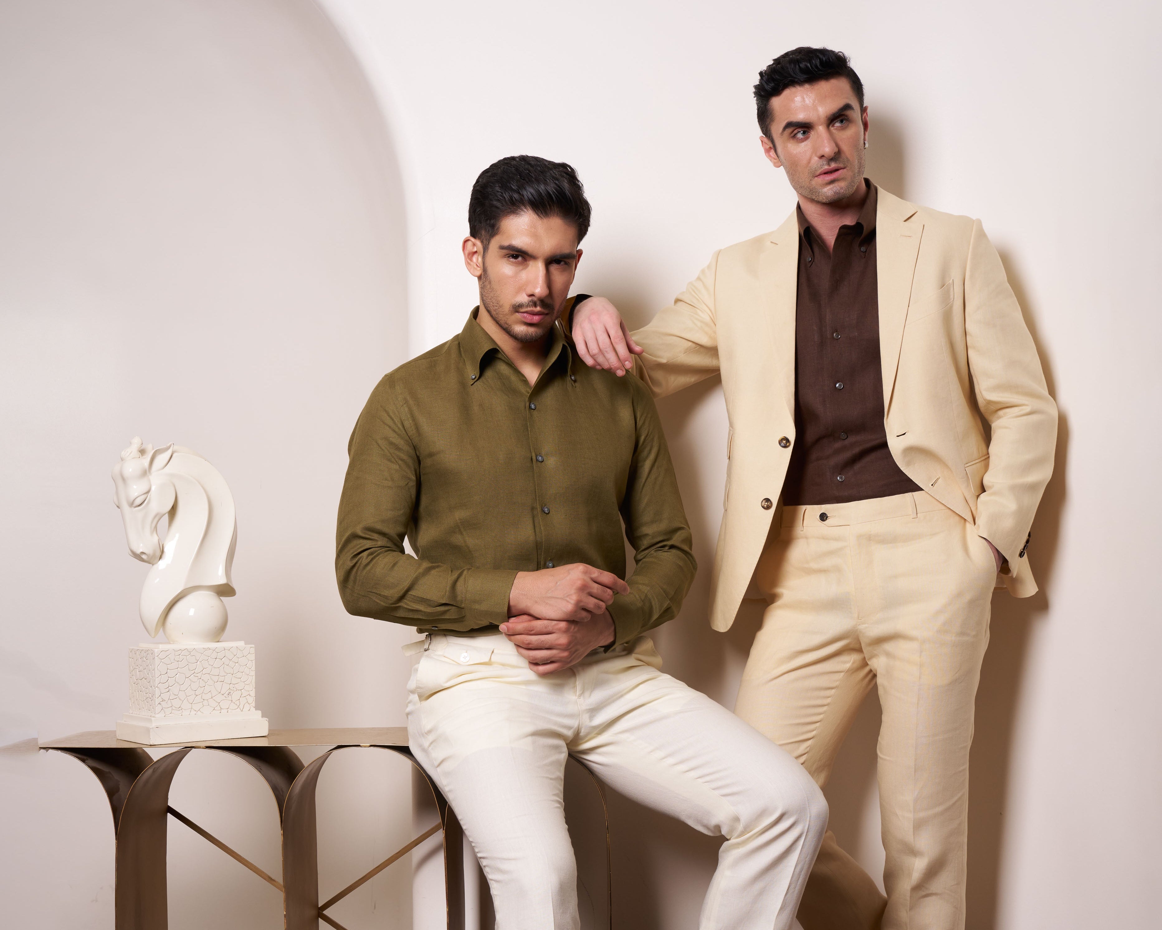 Two men in formal attire posing against a white wall with a decorative horse sculpture on a table.